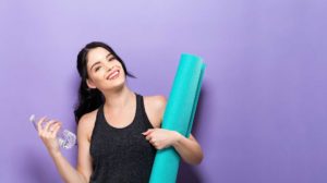 woman against purple background holding a yoga mat and a water bottle